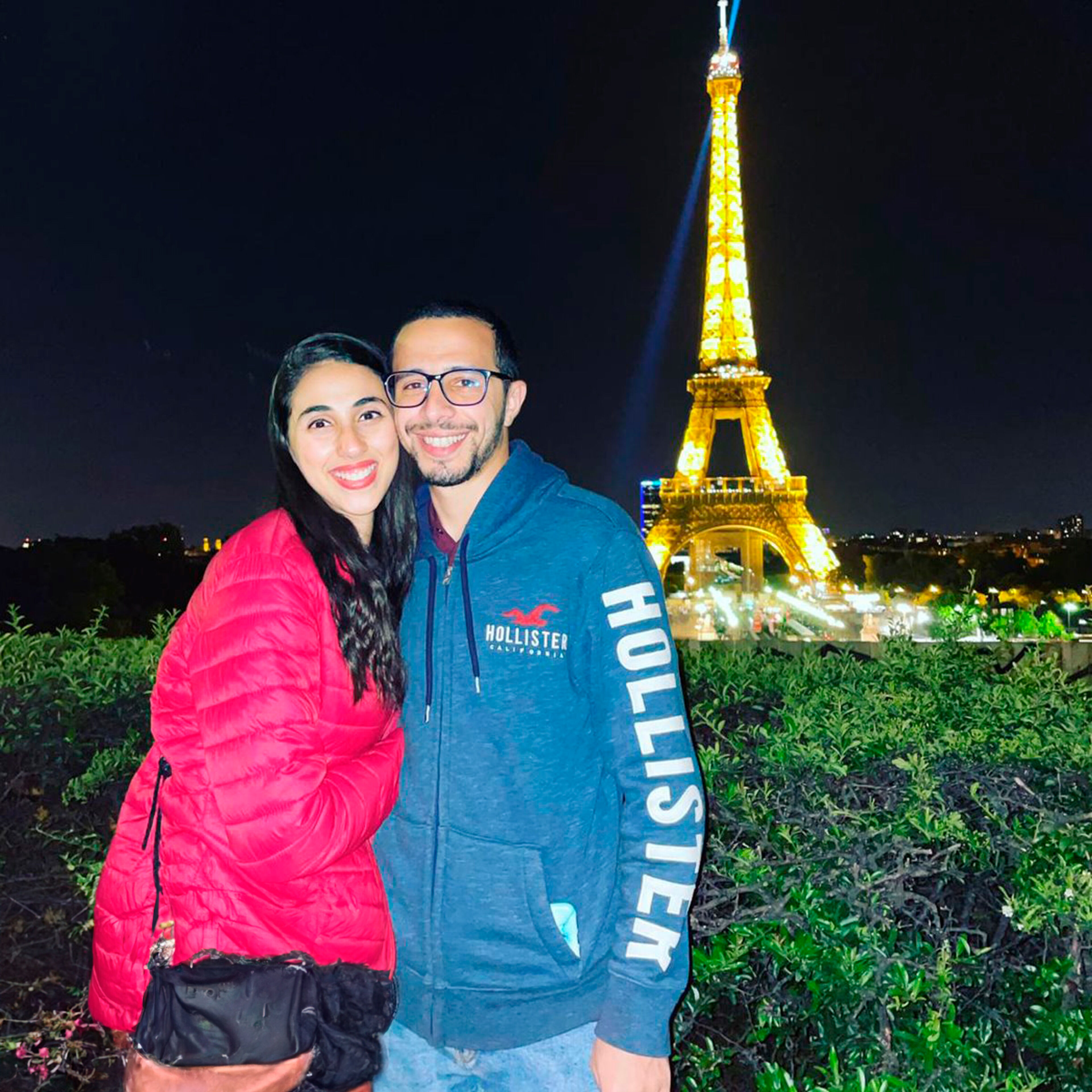 Un couple souriant pose pour une photo avec la Tour Eiffel illuminée en arrière-plan, mettant en valeur le monument emblématique de Paris la nuit.