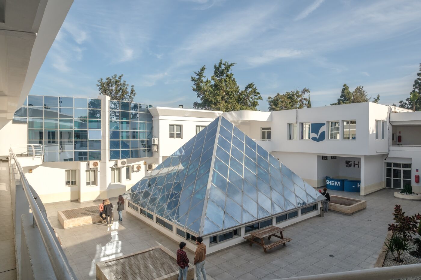 A contemporary school courtyard featuring a striking glass pyramid, reflective windows, and students socializing.