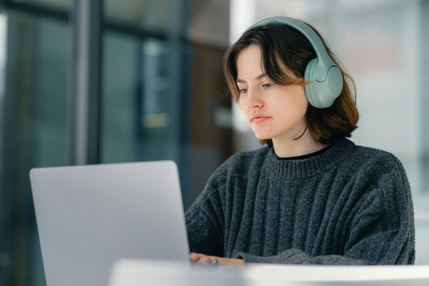 A young woman wearing green headphones is listening to music or a podcast while working on a grey laptop in a bright office setting.