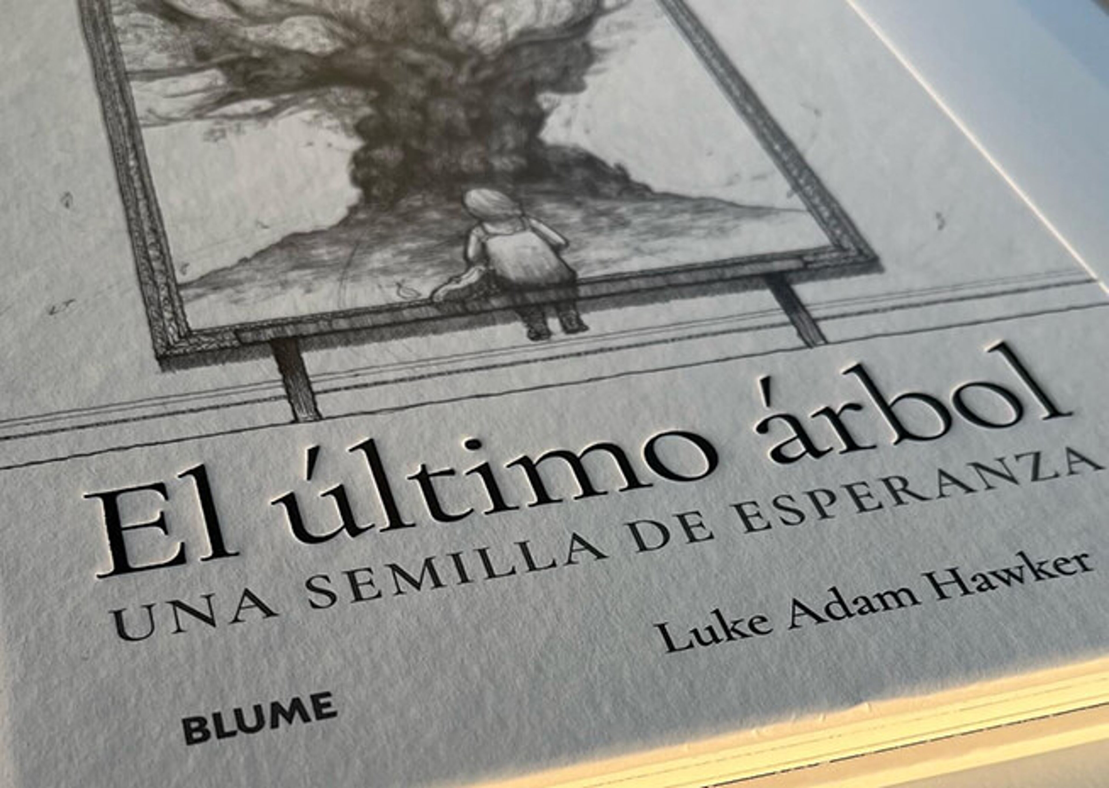 Detail of the book "El último árbol" featuring an illustration of a tree and a person, with the subtitle "UNA SEMILLA DE ESPERANZA" and the author's name, Luke Adam Hawker.