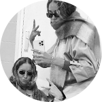 Black and white image of two young women wearing sunglasses and scarves, making peace signs with their hands.