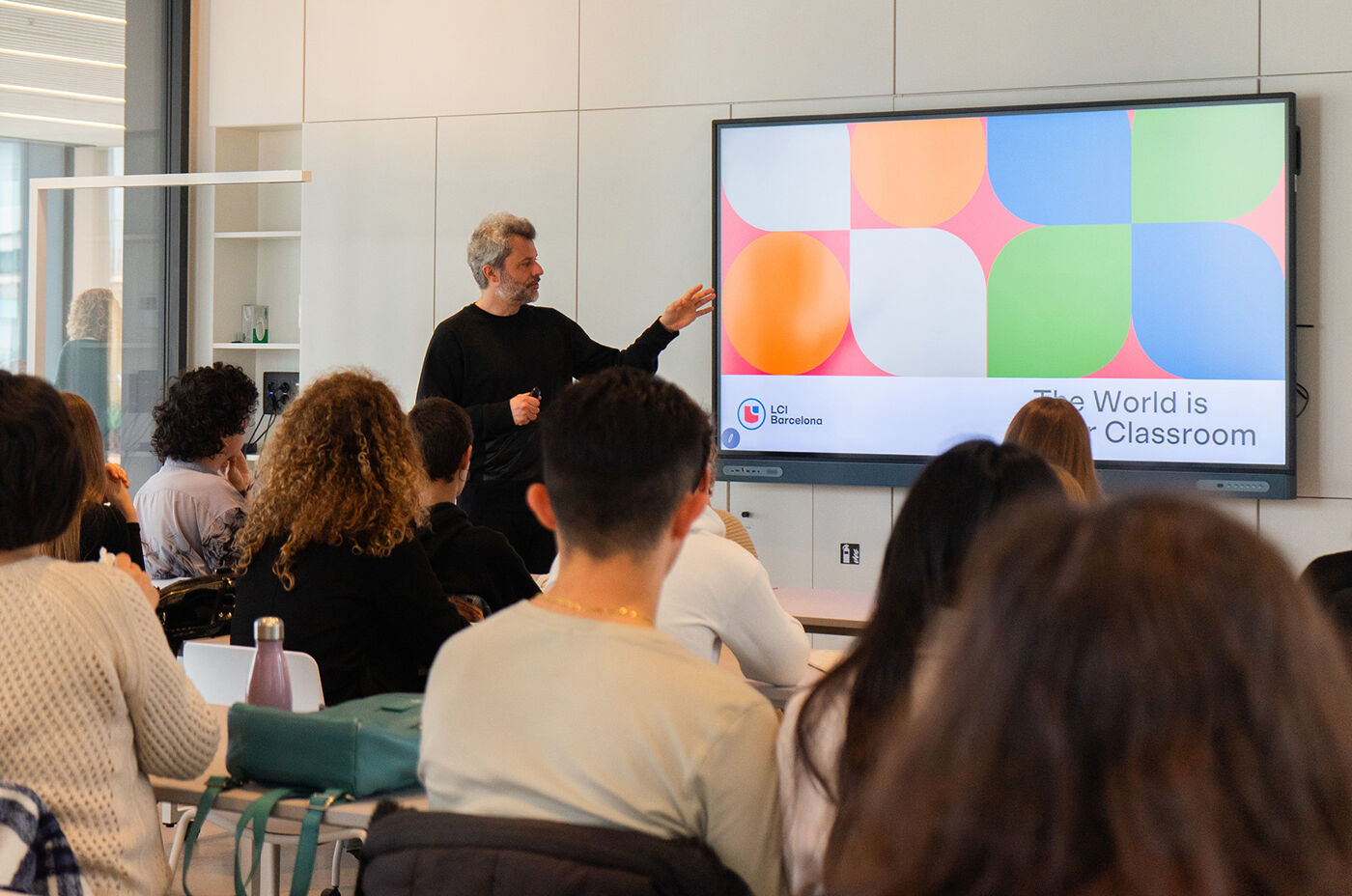 A man is presenting to a group of people in a classroom. He is standing in front of a screen that displays a colorful graphic and the words "The World Is Classroom."