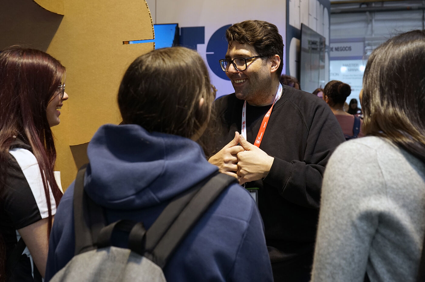 A group of people are gathered, seemingly engaged in a conversation. A man with glasses is the focal point, gesturing with his hands as he speaks to the others around him. The setting appears to be indoors, possibly at a conference or event.