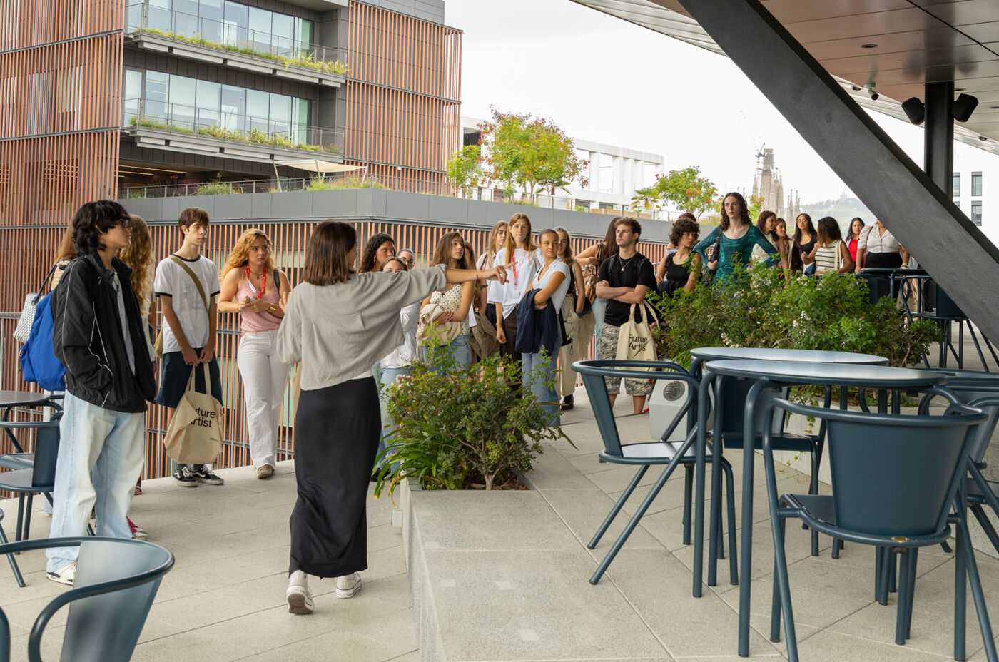 A group of people is gathered on a rooftop terrace, possibly a tour group, listening to a woman who appears to be their guide. The setting includes modern buildings and greenery.