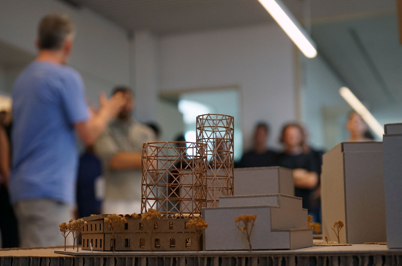 A group of people are gathered around a table looking at an architectural model. The model features cylindrical structures and rectangular buildings, suggesting a potential urban development project.
