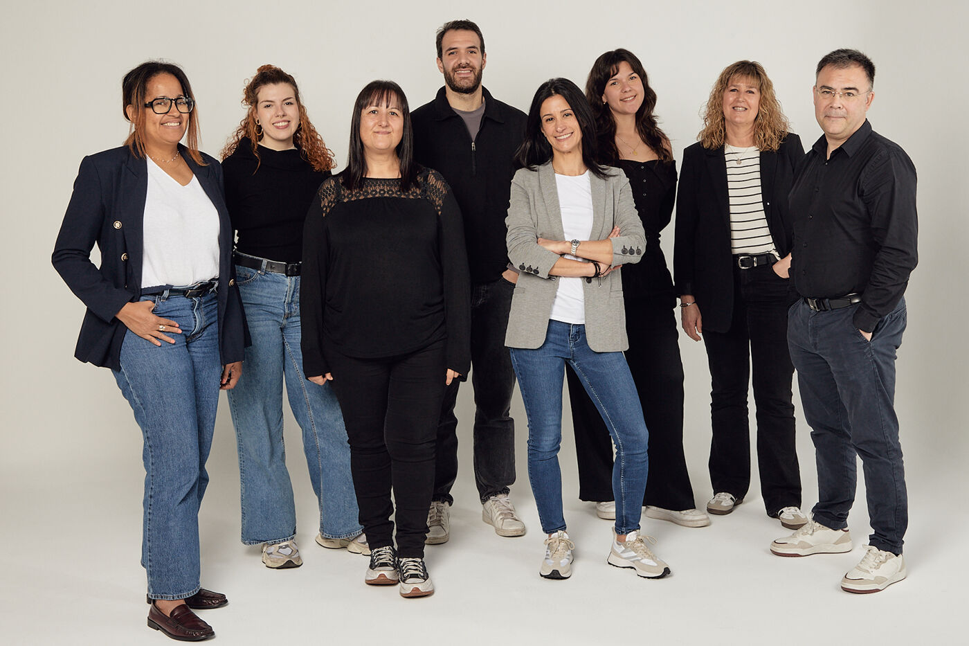 The image shows a group of nine people standing together in what appears to be a studio setting. They are dressed in a variety of casual and business-casual attire, and they are all facing the camera.