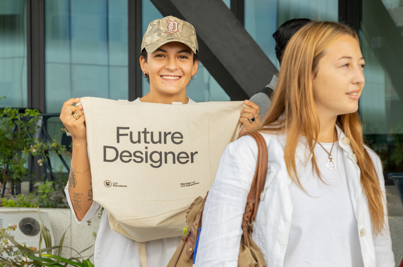 A person holding a tote bag that says "Future Designer" smiles at the camera. Another person with long hair stands to the side, looking away. Both are outside a building with large windows.