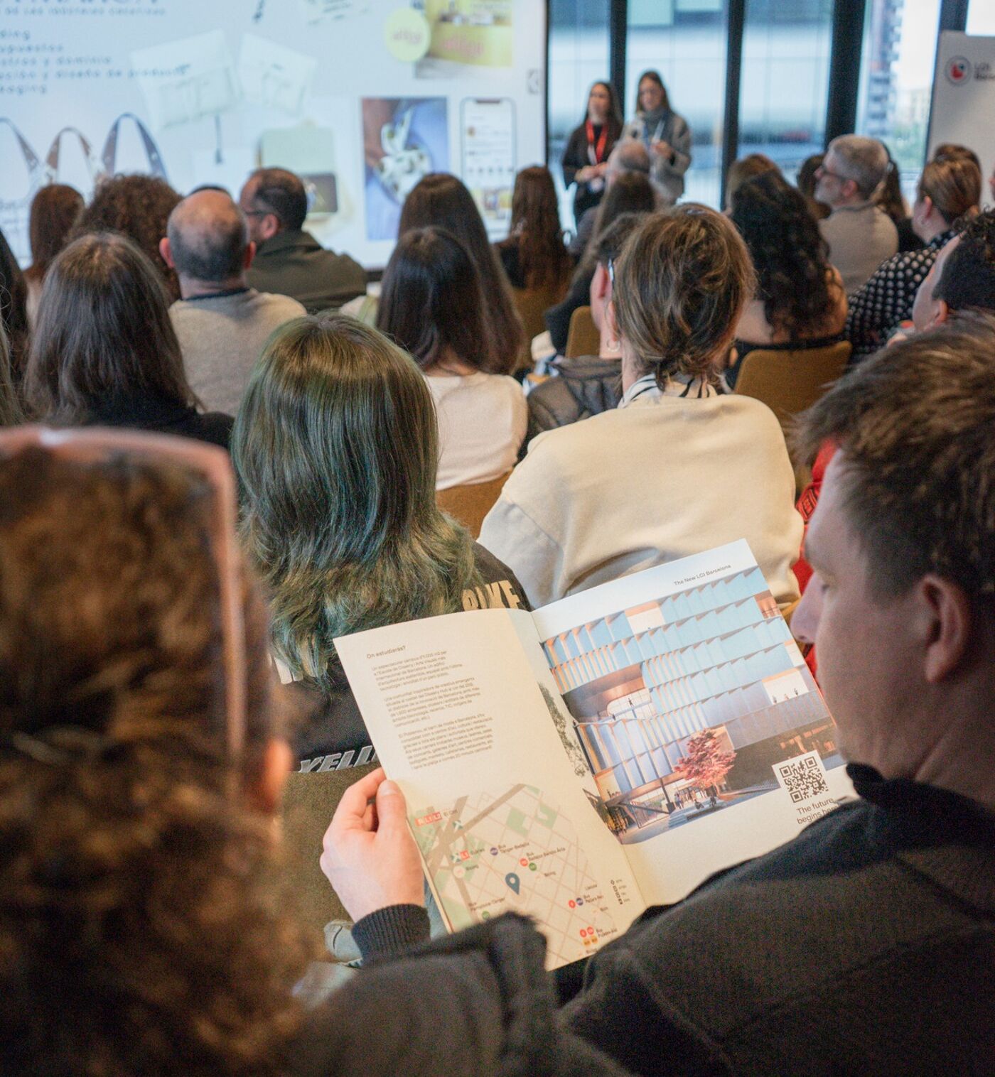 The image shows a conference or presentation with many people in attendance. Some individuals are looking at a magazine, while others are listening to the speakers.