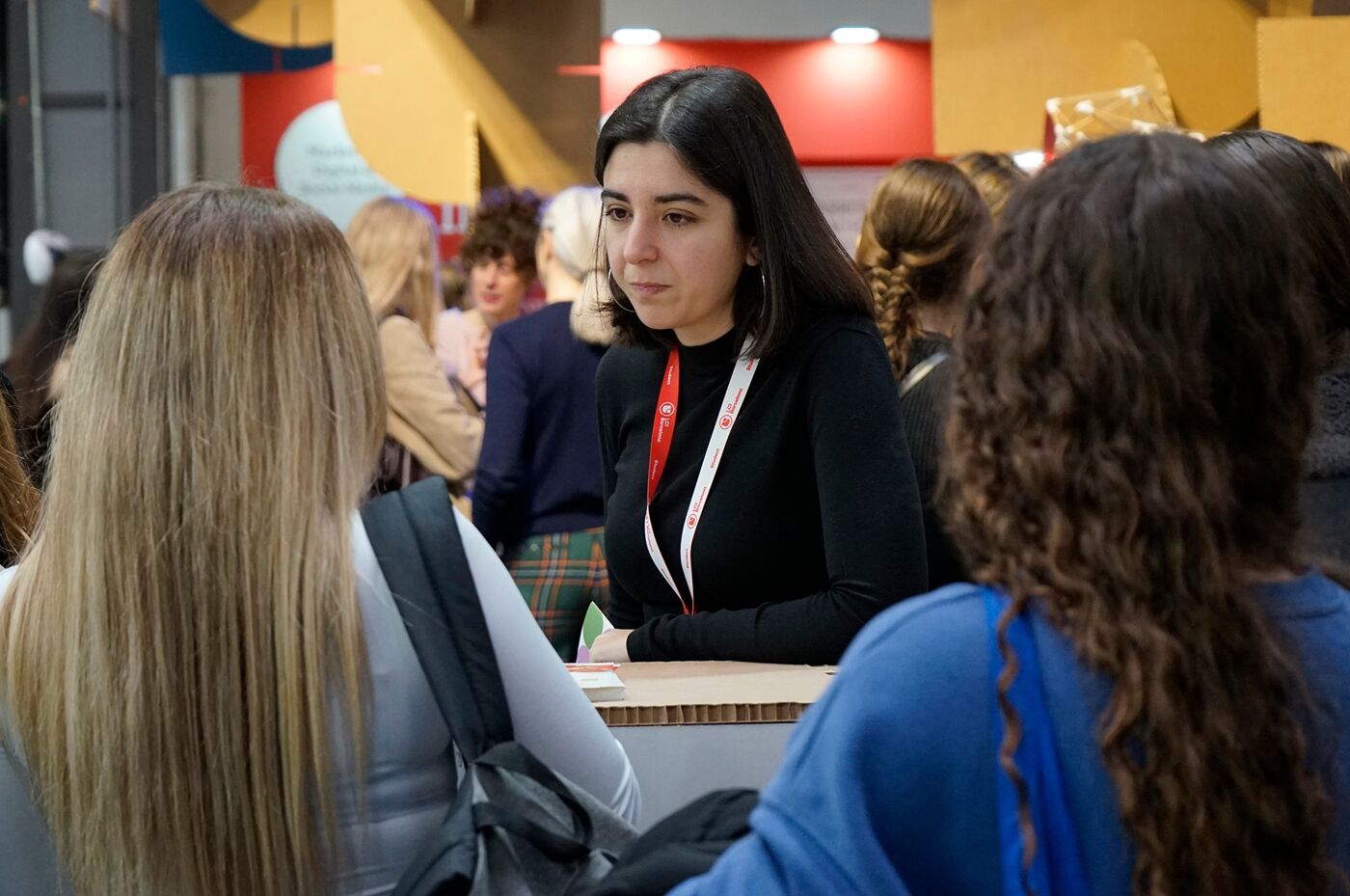 The image shows three women in a conversation. One woman is in the center, looking at the other two. They appear to be at an event or conference.