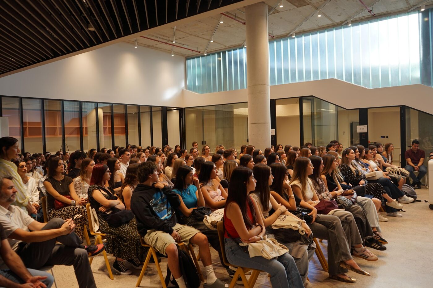 A large group of people, mostly young adults, attentively listens to a presentation in a spacious, modern hall. The setting features minimalist design elements, large windows, and a high ceiling. Attendees are seated on wooden chairs, suggesting an educational or professional event.