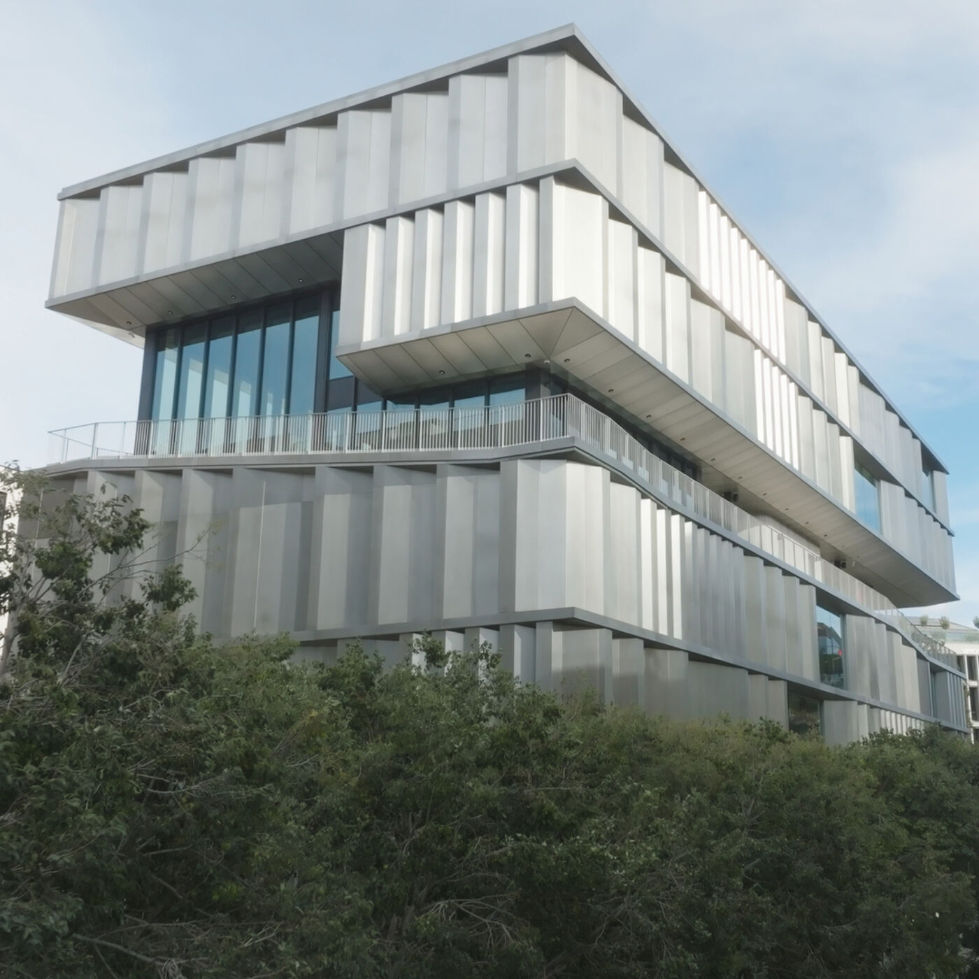 A contemporary building featuring a unique tiered design and sleek metallic facade, surrounded by greenery. The structure's large glass windows and layered appearance reflect a modern architectural style, likely serving as an office, gallery, or educational facility.