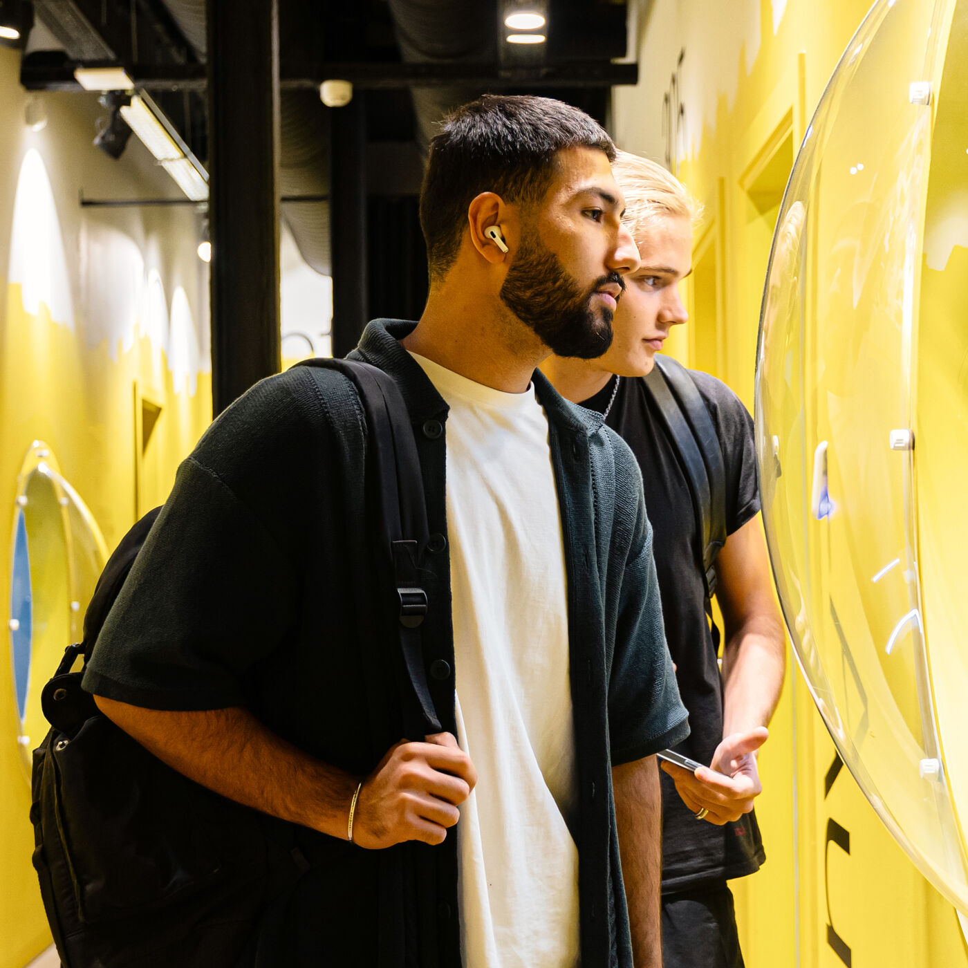  Two young men, both carrying backpacks, closely examine a display in a brightly lit yellow room. One wears wireless earbuds, and both appear engaged and focused. Their casual attire suggests they may be students or tourists exploring an exhibit or store.