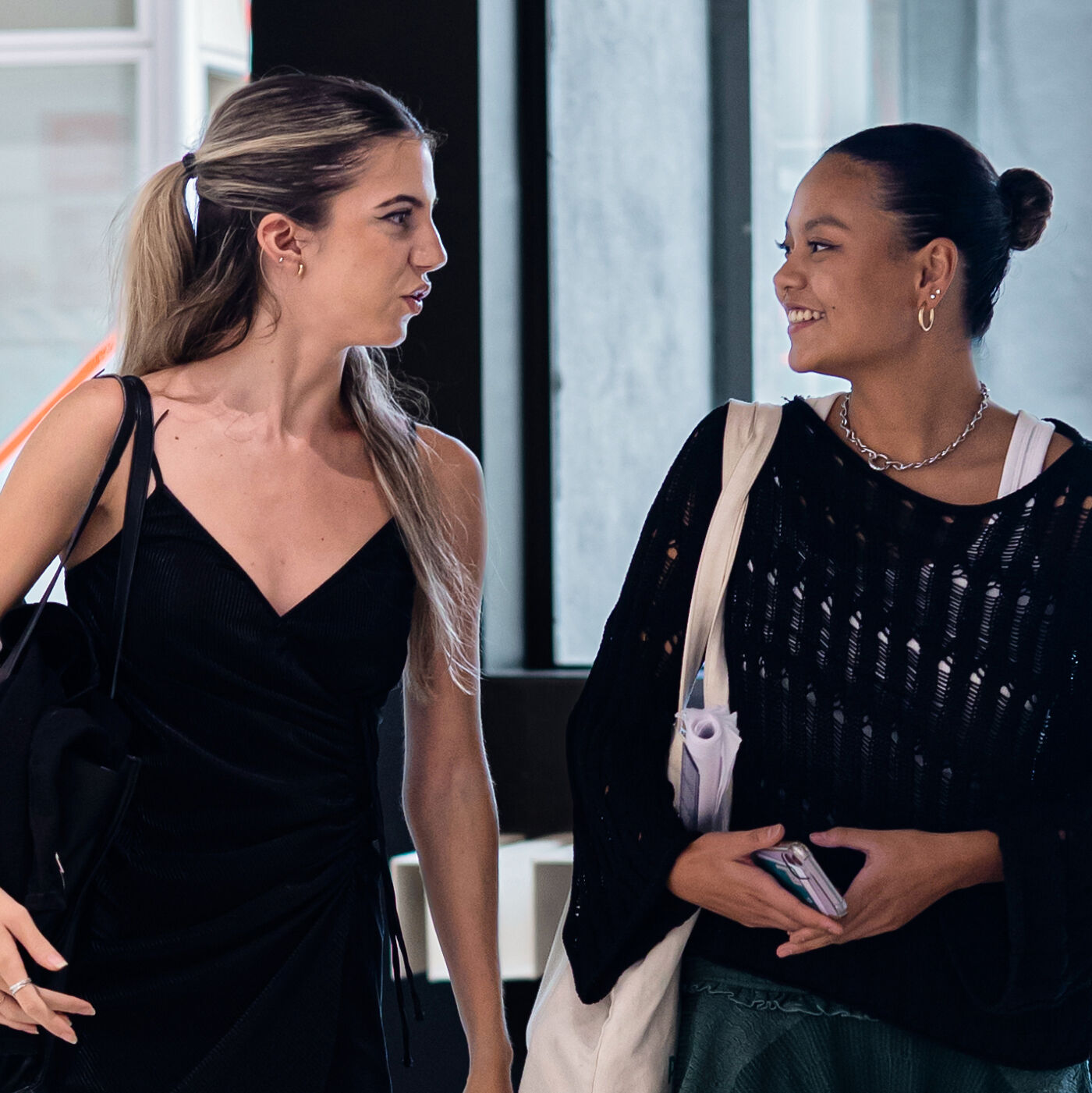 Two young women are walking together, engaging in a lively conversation. One wears a black dress and ponytail, while the other is dressed in a black knit top with a bun hairstyle. Both carry bags, suggesting they are either students or young professionals.