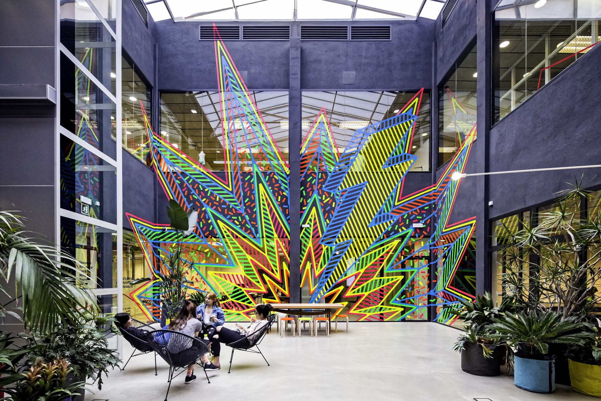 People relax in an atrium featuring a vibrant, geometric art installation surrounded by lush greenery.