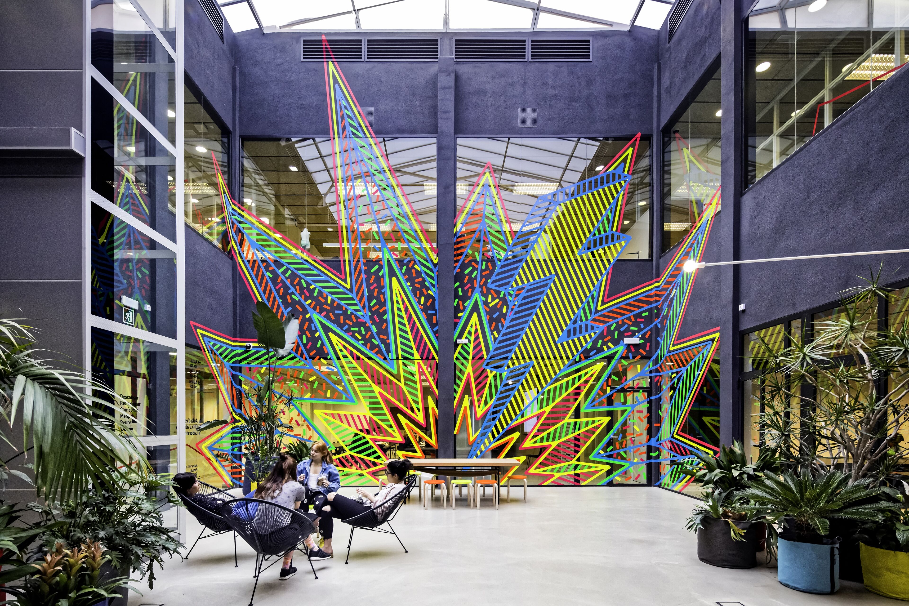 People relax in an atrium featuring a vibrant, geometric art installation surrounded by lush greenery.