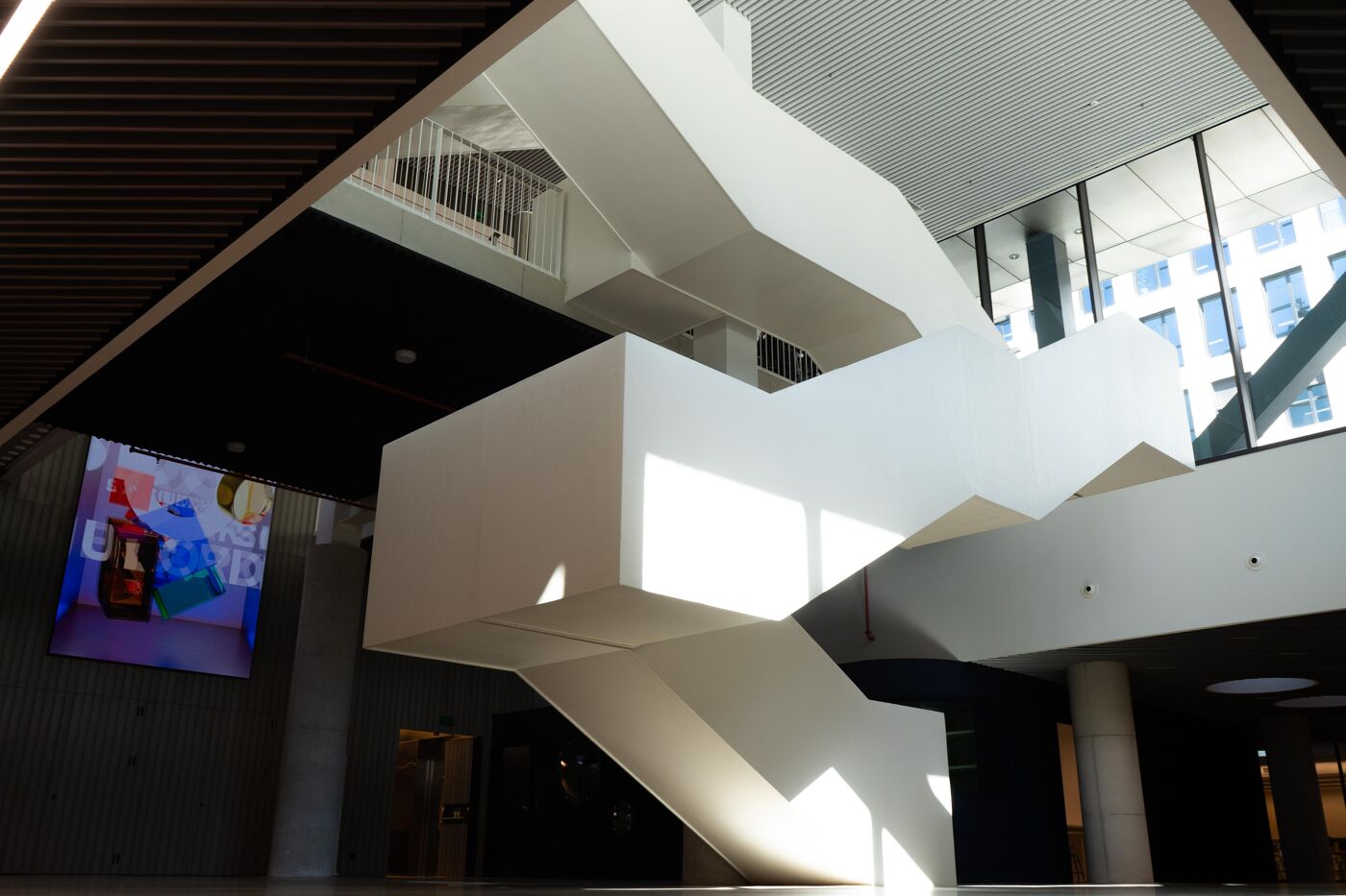 This image shows a modern indoor staircase with a unique, geometric structure. The stark white design contrasts with surrounding shadows, while large windows provide natural light, adding depth to the minimalist architectural style.