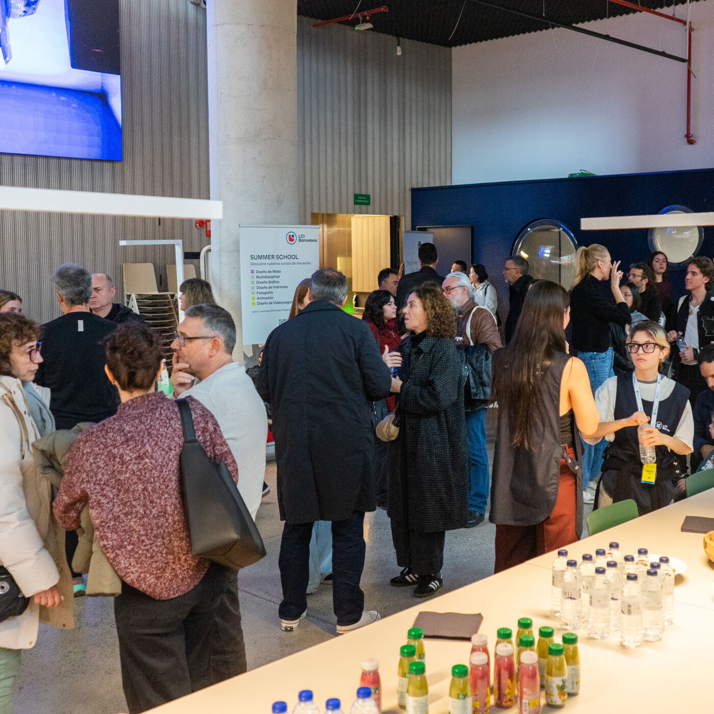 A group of people are gathered in a large, open space, possibly for an event or conference. Some are standing and talking, while others are seated. There's a table with drinks in the foreground.