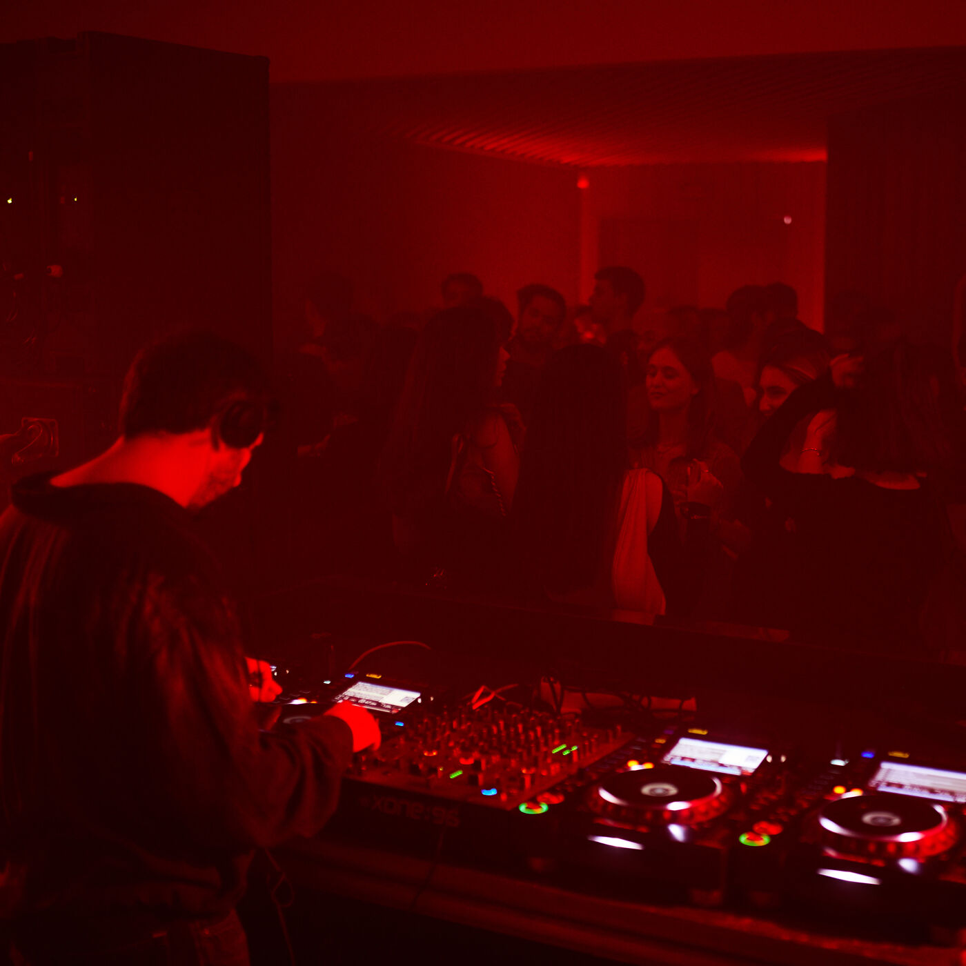 The image shows a DJ performing in a dimly lit club with red lighting. The DJ is facing away from the camera, focused on the mixing equipment. A crowd of people can be seen in the background.