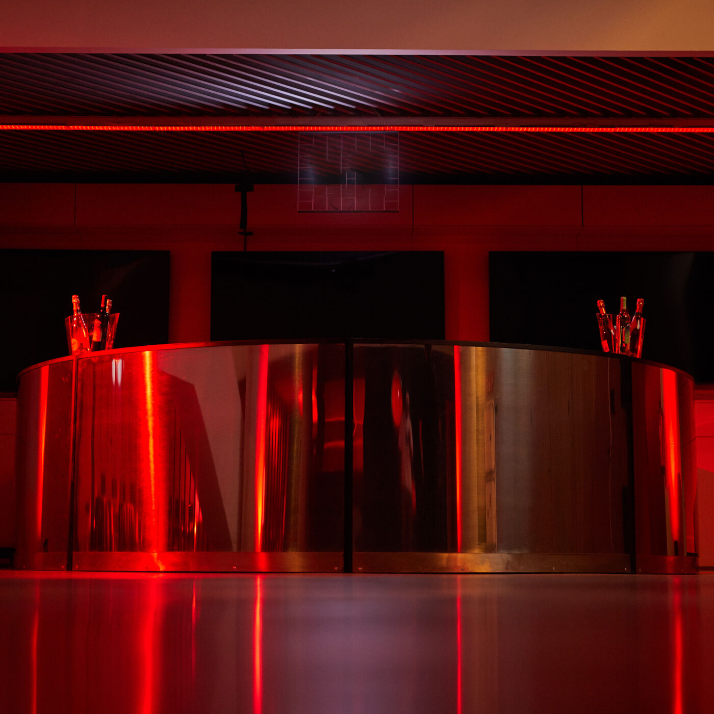 The image shows a curved, metallic bar illuminated by red lighting. The bar appears to be in a dimly lit setting, with reflections visible on its surface. Glasses are placed on the bar's ends.