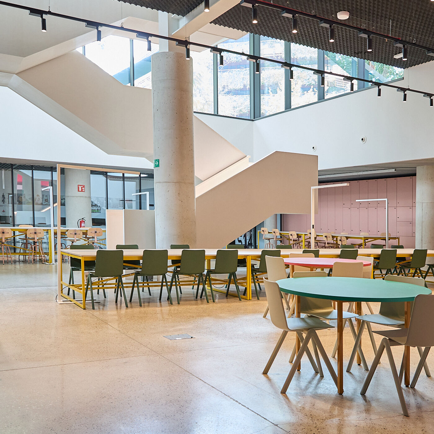 The image shows a brightly lit, modern interior space with tables and chairs arranged for dining or working. A large concrete pillar and staircase are prominent features, with natural light streaming in from above.