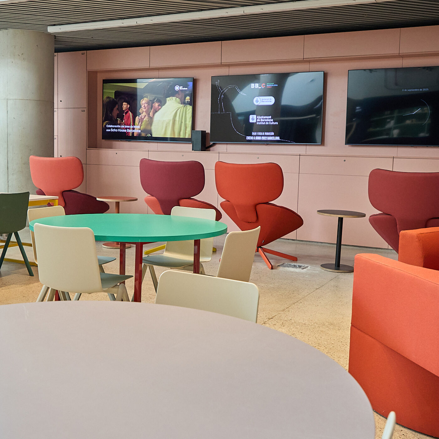 The image shows a modern lounge area with colorful chairs and tables. There are three TVs mounted on the wall, and the overall ambiance is bright and inviting.
