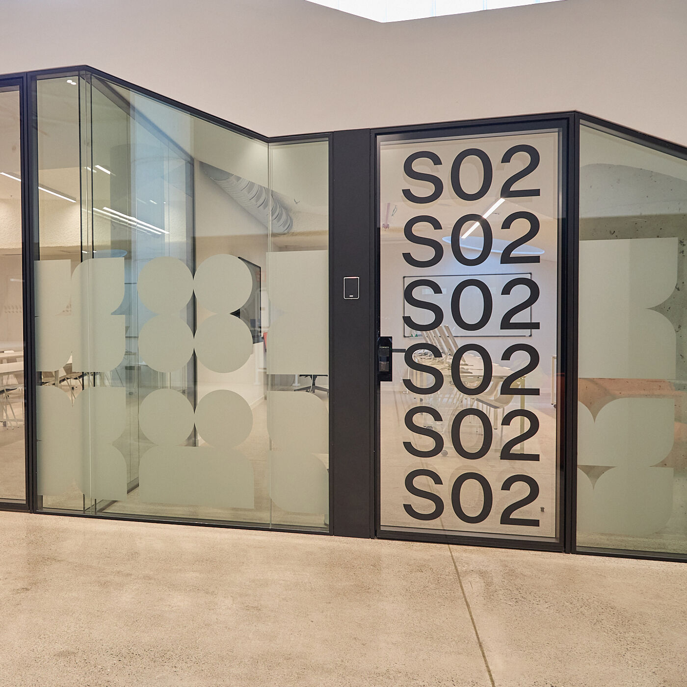 The image shows a modern office space with glass-walled rooms. The door to one room is labeled "SO2" multiple times vertically. The walls have frosted designs, and the overall aesthetic is clean and contemporary.