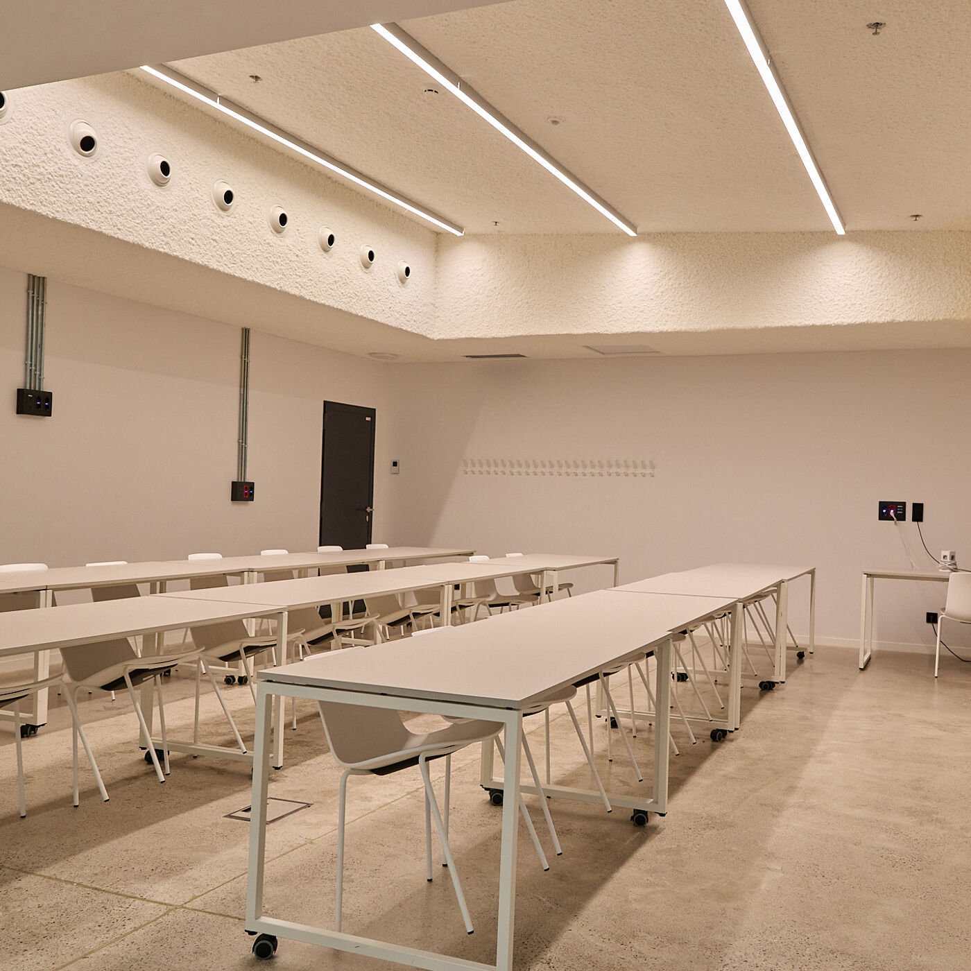 The image shows a bright, modern classroom with rows of tables and chairs. The room has a minimalist design with white walls and ceiling, and long fluorescent lights. There's a door on the left side of the room.