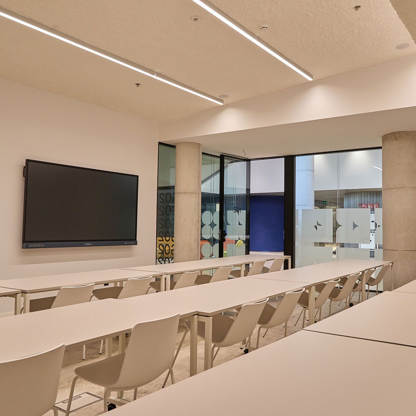 The image shows a well-lit, modern conference room with long tables and chairs arranged in rows. A large screen is mounted on the wall, and the room features large windows and minimalist decor.