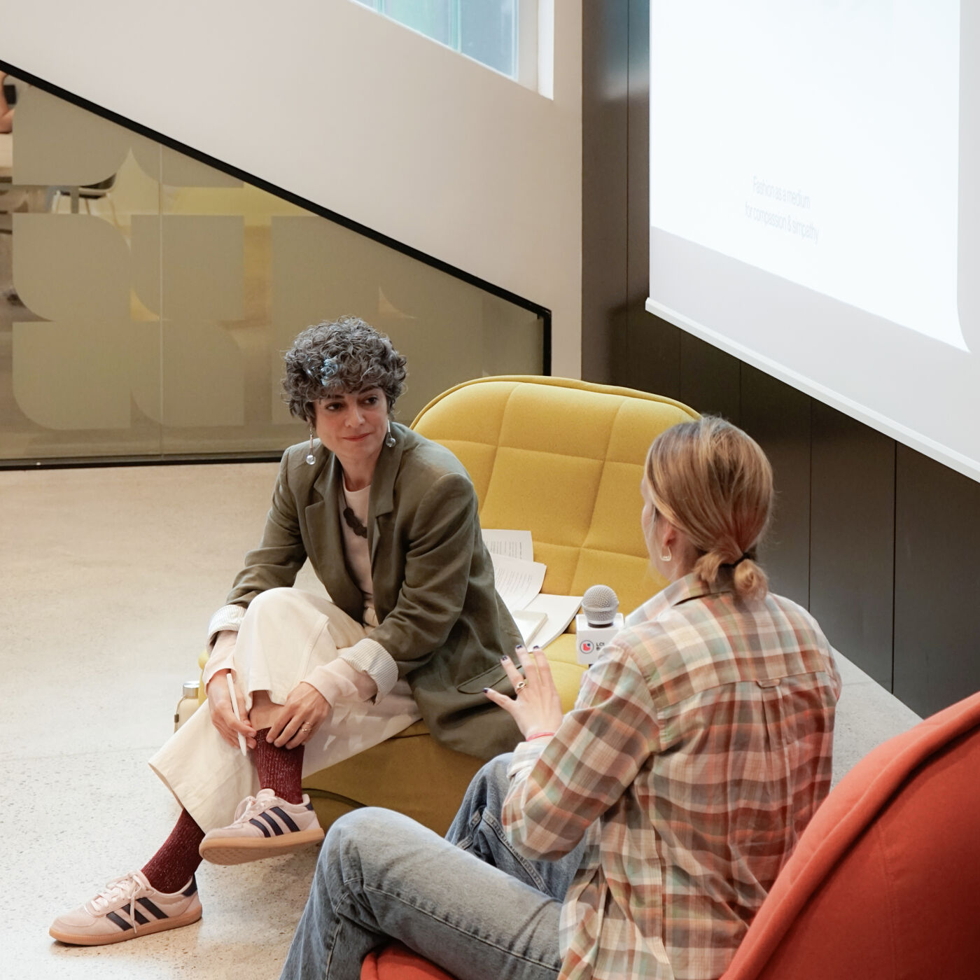 Two women are sitting and talking in a modern, bright space. One woman is wearing a blazer and sneakers, while the other is in a plaid shirt. A microphone is present, suggesting a possible interview or discussion.