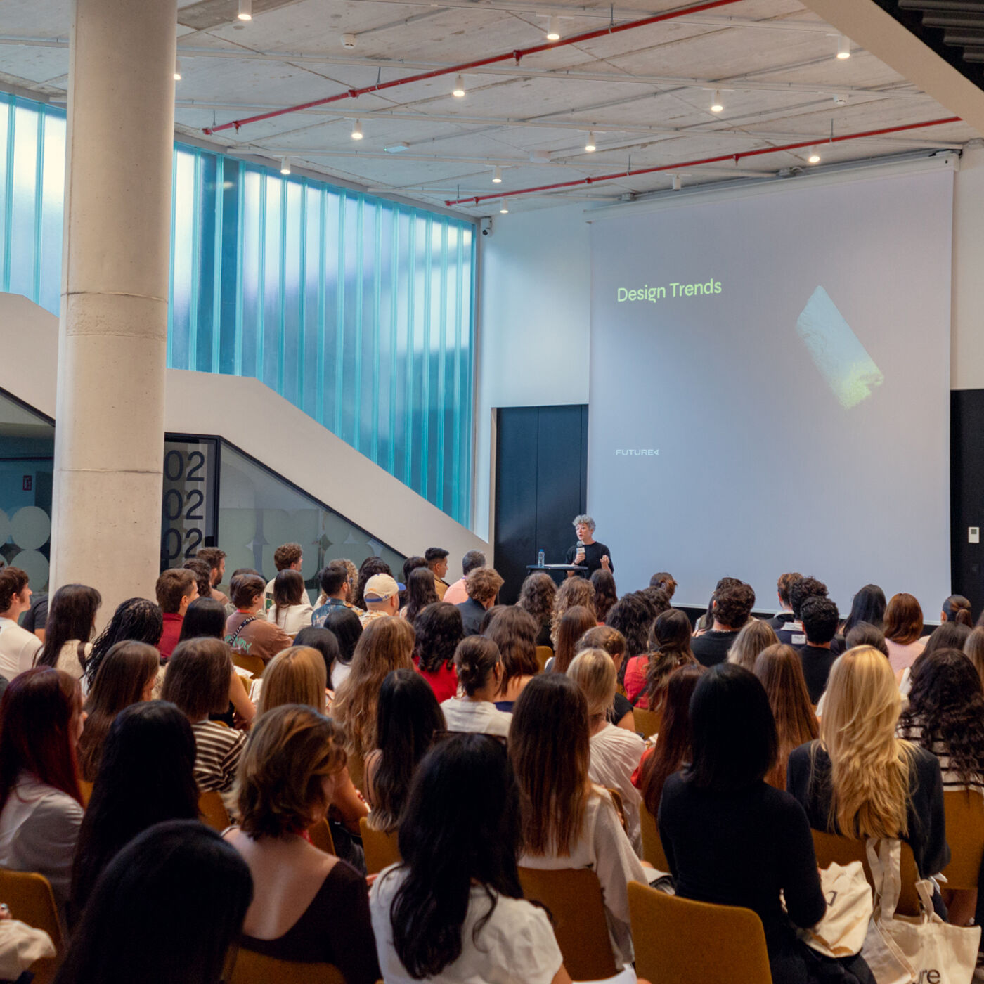 A large group of people are seated in an auditorium, facing a presenter who is standing on a stage. A screen displays the words "Design Trends" and an image. The room has a modern architectural design.