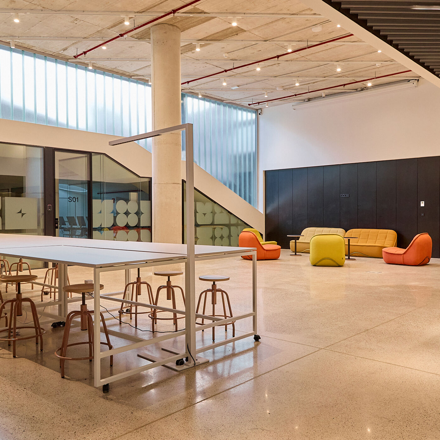 The image shows a modern, open-plan office space with a large table in the foreground, surrounded by stools. In the background, there are sofas and chairs, suggesting a collaborative or lounge area. The space is well-lit with natural light from large windows.