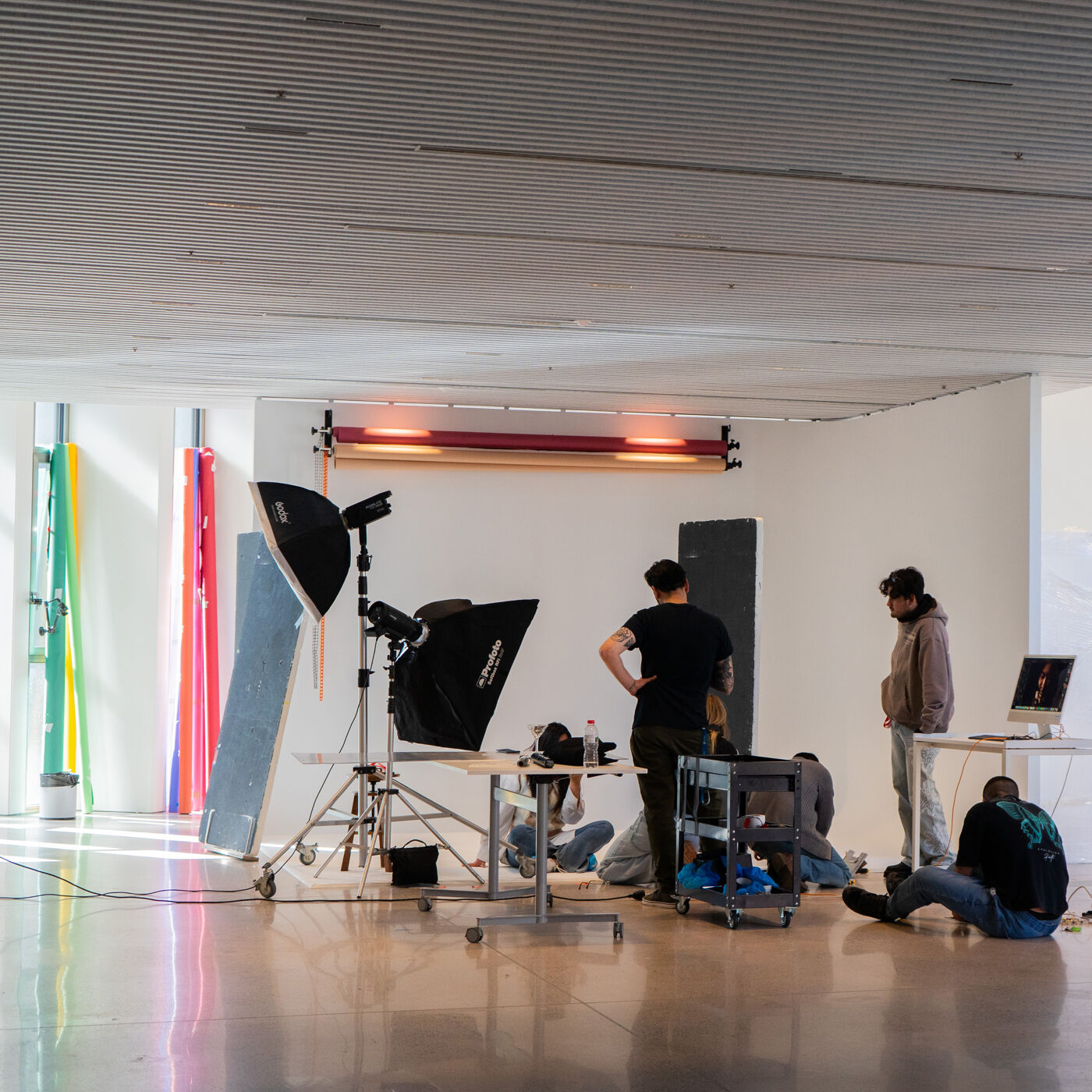 A photo shoot is in progress in a studio with lighting equipment, backdrops, and several people. Some individuals are standing, while others are seated or working on the set.