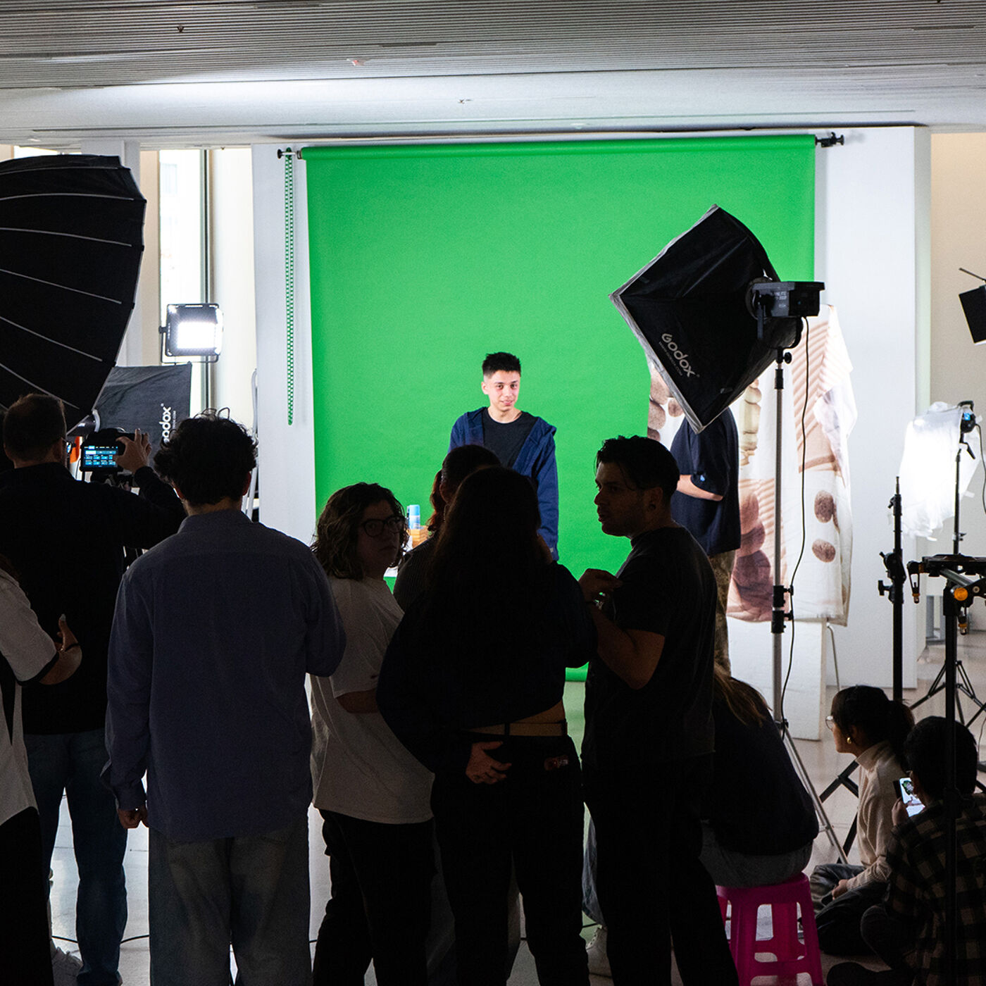 A group of people are gathered around a person standing in front of a green screen. There are lights and camera equipment set up, suggesting a photoshoot or video shoot is in progress.