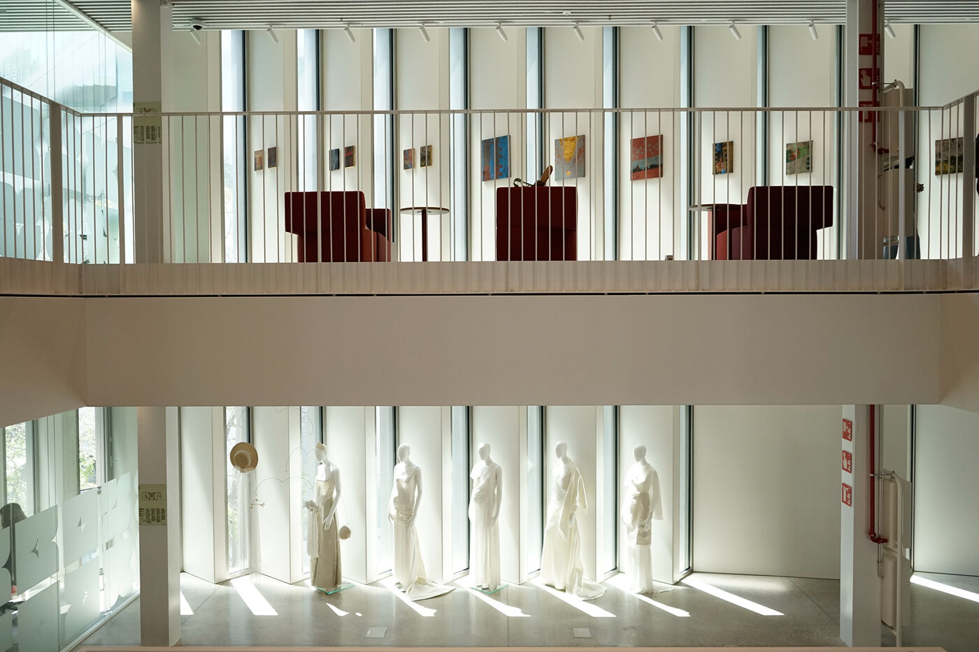 The image shows a multi-level interior space with mannequins dressed in white garments on the lower level. The upper level features seating areas and artwork displayed against a backdrop of large windows.