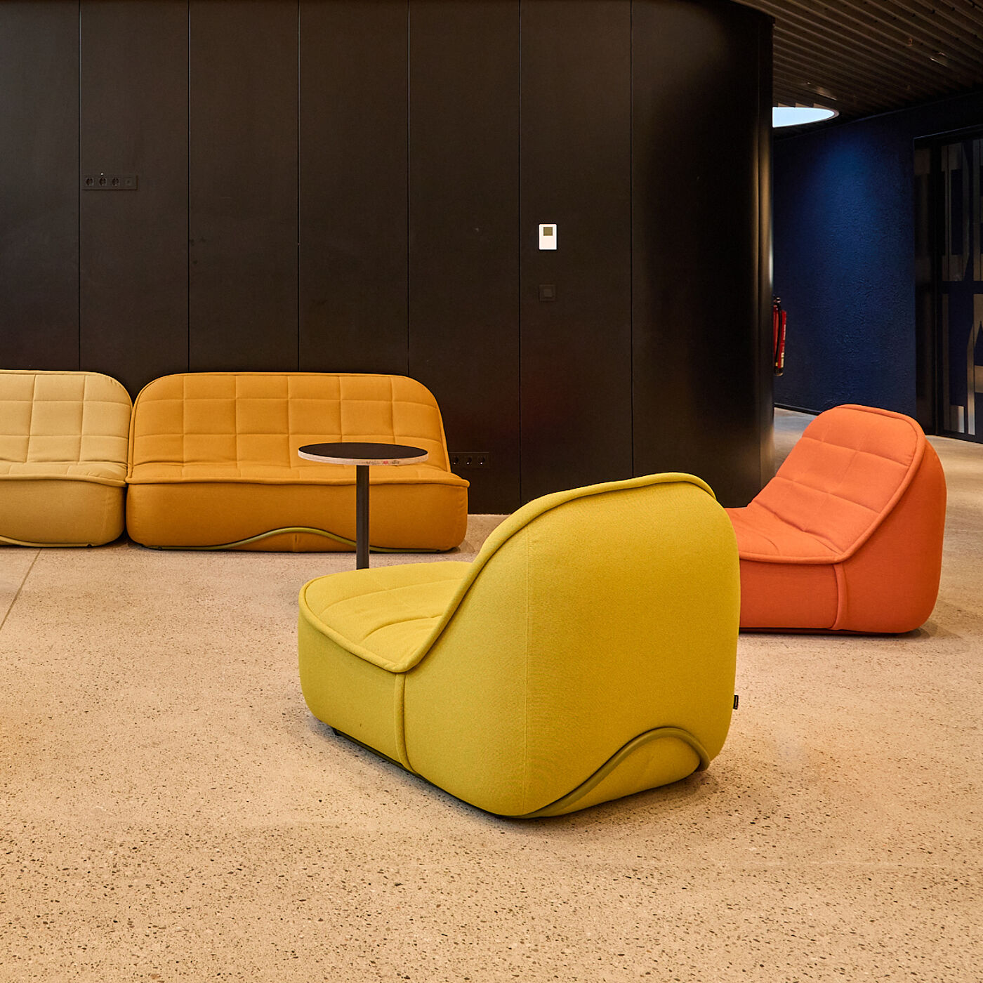 The image shows a modern lounge area with colorful, modular seating. There are yellow, orange, and mustard-colored chairs and a small black table on a speckled floor against a dark wall.