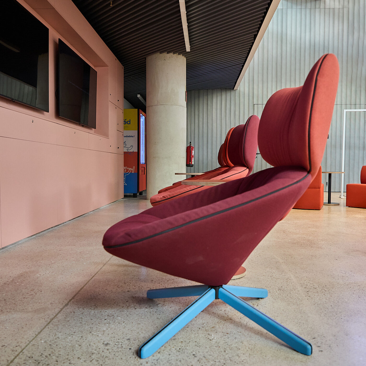 The image shows a row of modern, red chairs with blue bases, likely in a waiting area or lounge. The chairs have a unique, angled design and are positioned against a wall with a pinkish hue.