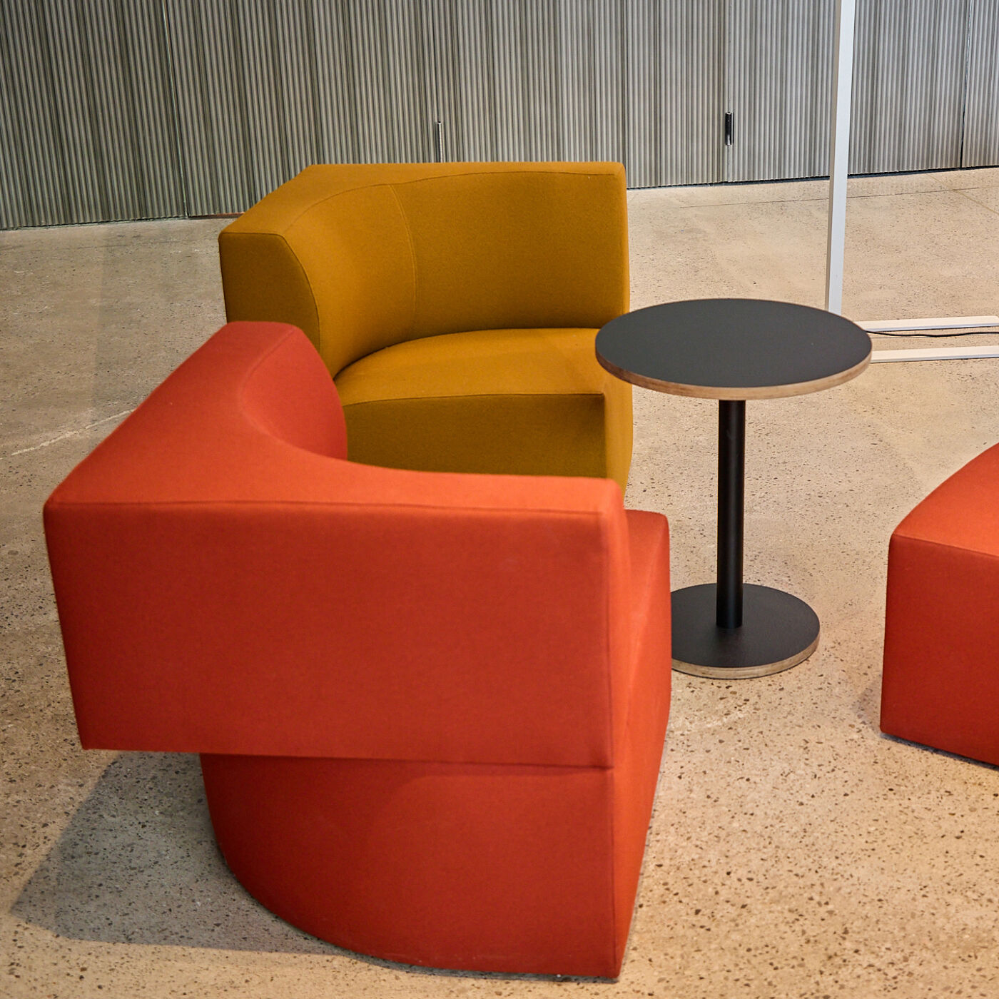The image shows a modern lounge area with curved, colorful armchairs in orange and yellow, arranged around a small, round black table. The setting suggests a contemporary and inviting space.