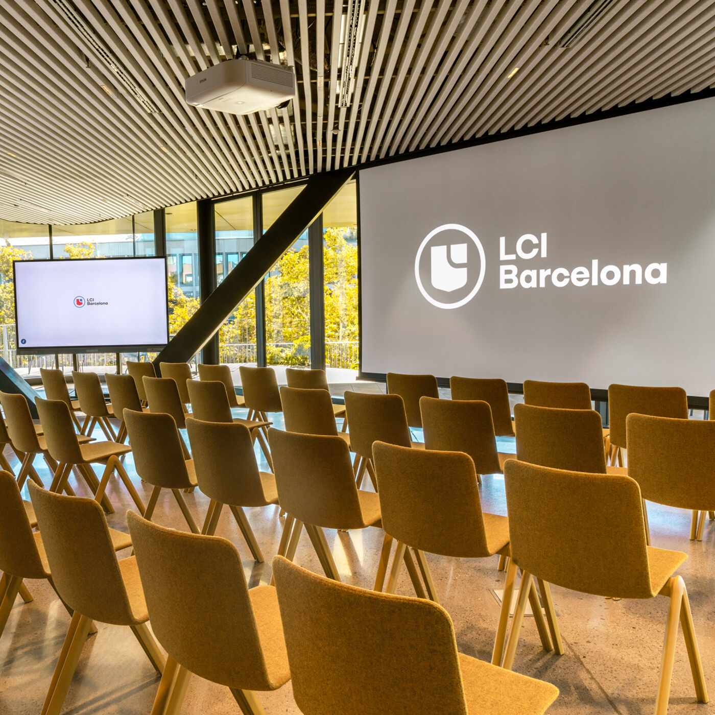 The image shows a modern conference room with rows of yellow chairs facing a large screen displaying the LCI Barcelona logo. Natural light streams in through the windows, illuminating the room's sleek design.
