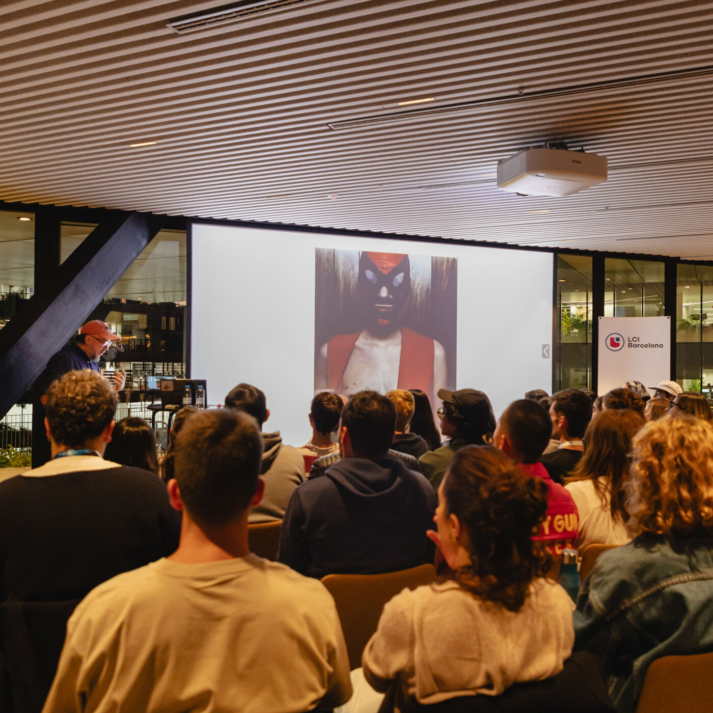 A group of people are watching a presentation on a screen. The screen displays an image of a person wearing a red mask. The audience is seated in rows, facing the screen. A presenter is standing to the left of the screen.