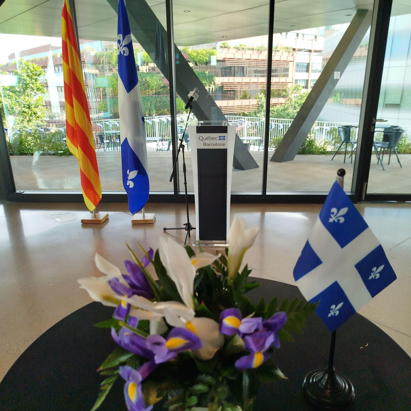 The image shows a room with large windows, featuring three flags: one with yellow and red stripes, one with a blue and white fleur-de-lis, and a smaller Quebec flag on a stand. A flower arrangement sits in the foreground.