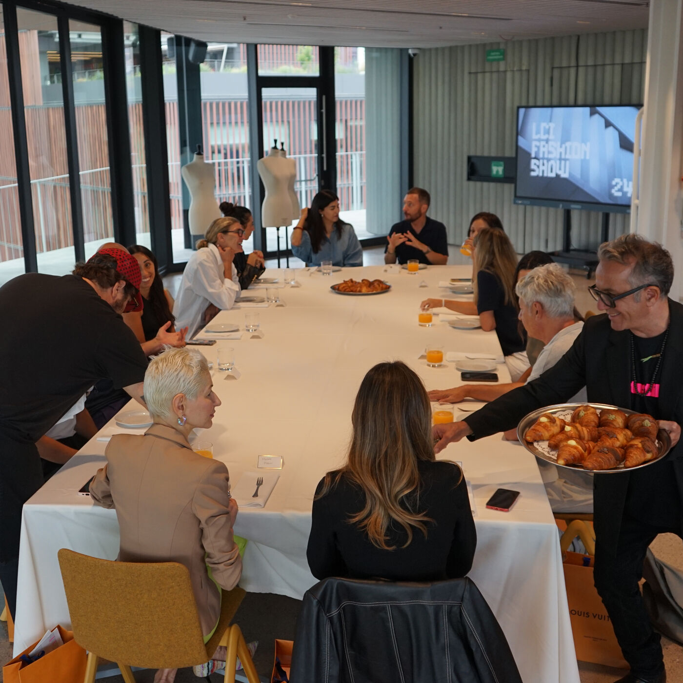 A group of people are gathered around a long table, possibly for a meeting or a meal. There's a TV screen in the background displaying 'Let Fashion Show'. A person is serving food from a tray.