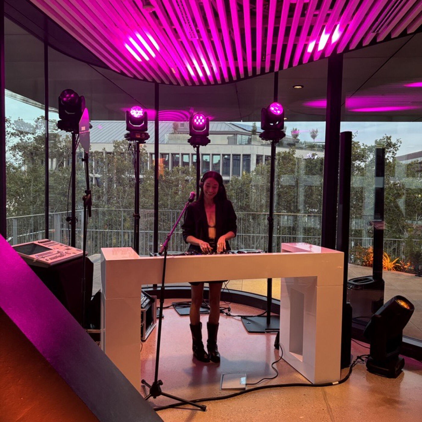 A woman stands behind a white DJ booth, surrounded by lighting equipment, in a room with large windows overlooking an outdoor area. The room is illuminated with pink and purple lights, creating a vibrant atmosphere.