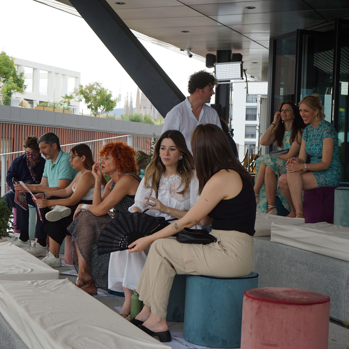 A group of people are gathered on a rooftop patio. Some are seated on benches and stools, while others are standing. They appear to be engaged in conversation or watching something. The setting suggests a social event or gathering.