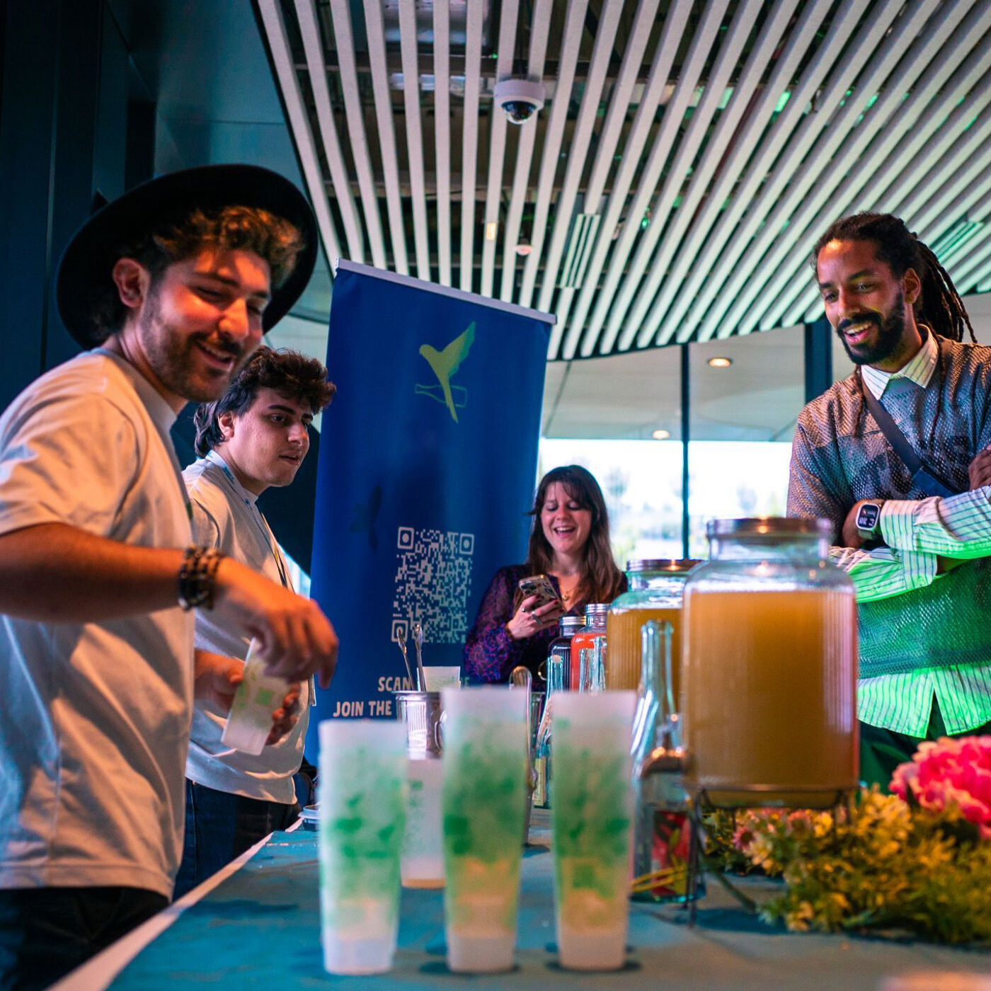 The image shows a group of people standing around a table with drinks. There is a banner in the background. The people appear to be at an event or gathering, possibly related to the banner's theme.