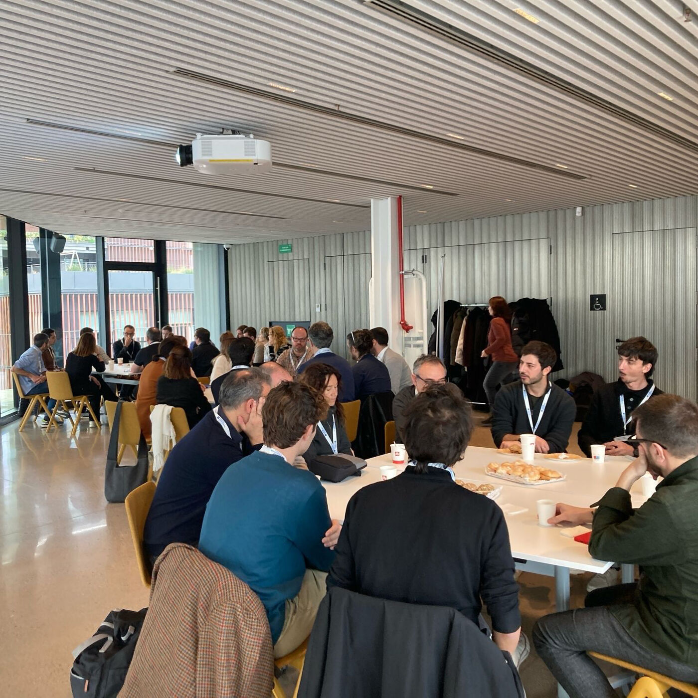The image shows a group of people gathered around tables in a well-lit room, possibly a conference or meeting space. They appear to be engaged in conversations, with some individuals looking at each other and others looking down at the table.