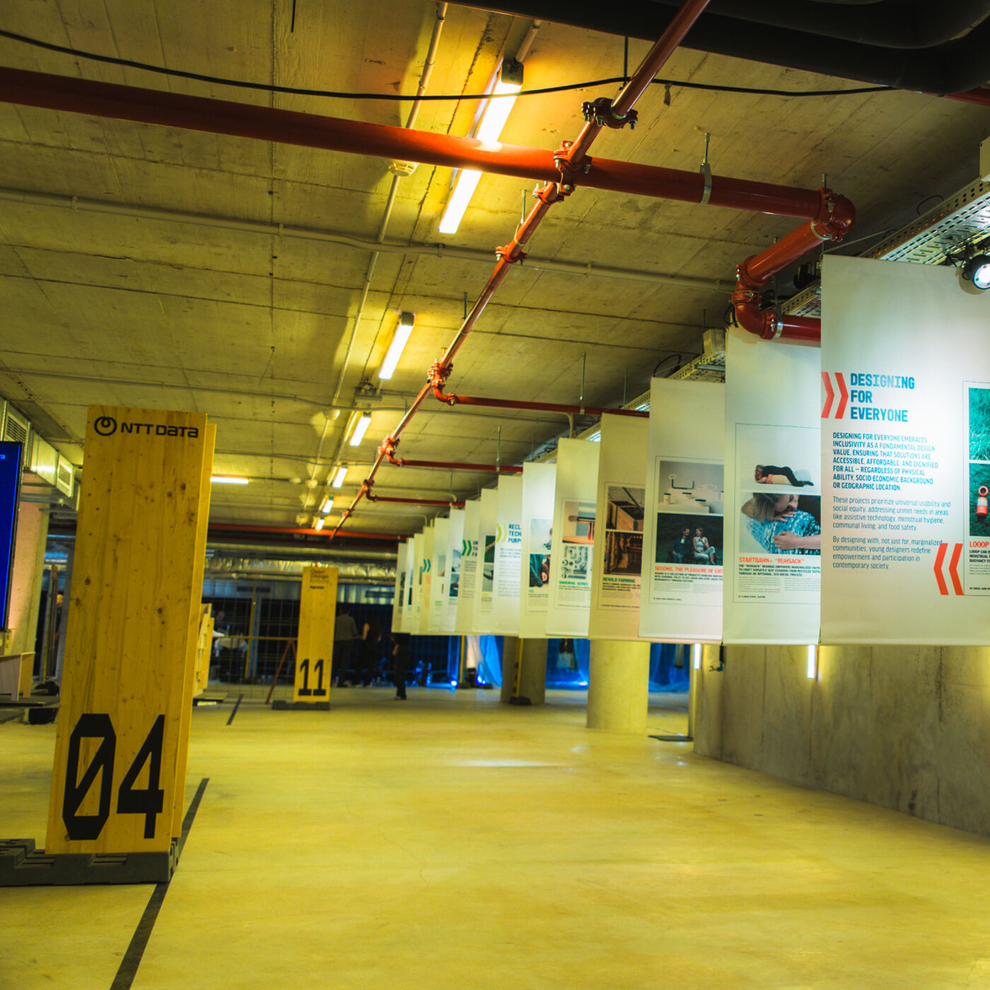 The image shows an indoor space with a low ceiling and exposed pipes. There are banners hanging from the ceiling, and a wooden structure with the number 04 is visible on the left. The floor is light-colored and reflective.