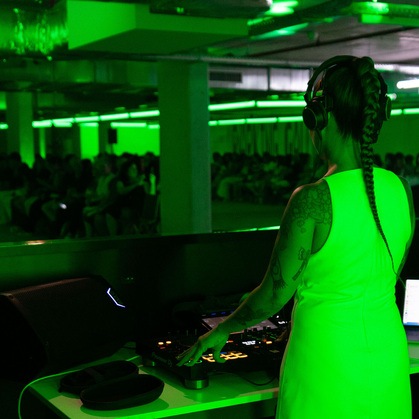 A woman with a braid is DJing in a green-lit room. She is wearing a neon green dress and headphones, standing behind a DJ console with a crowd in the background.