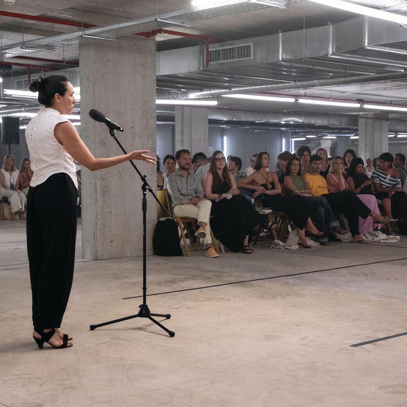 A woman is speaking into a microphone in front of a seated audience. The setting appears to be an indoor event space with concrete pillars and visible ceiling infrastructure. People are attentively listening to the speaker.