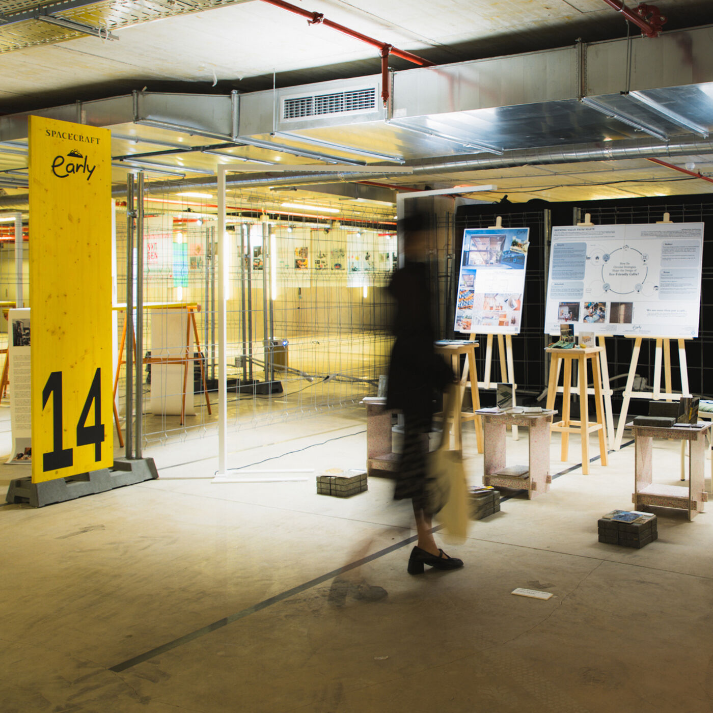 The image shows an indoor exhibition space with posters and displays. A person is walking through the space, and there is a yellow sign with the number 14 on it. The area appears to be a repurposed or temporary exhibition venue.