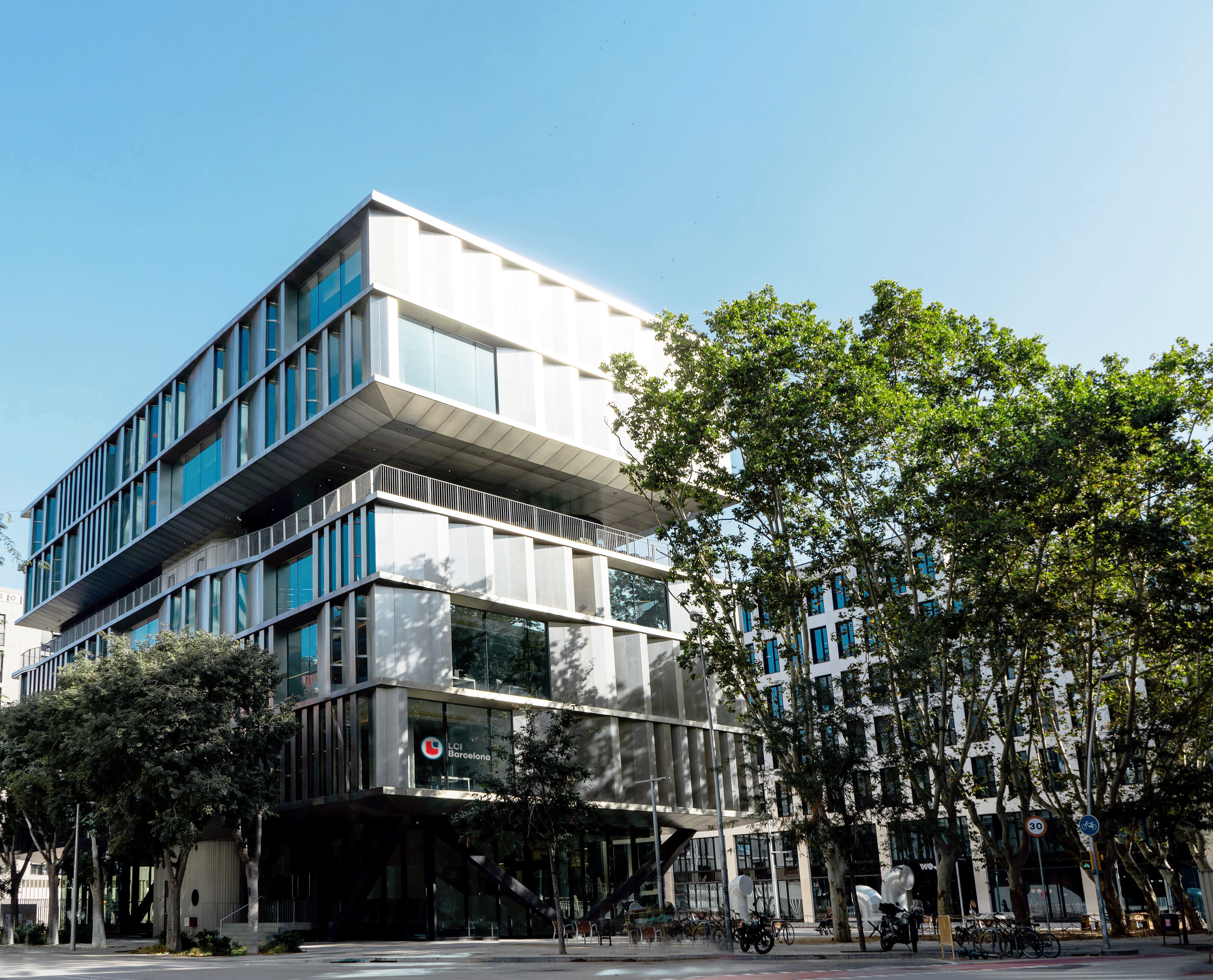 A modern, multi-story building with a unique architectural design stands amidst lush green trees under a clear blue sky.